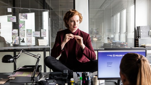 Linda Selb (Luise Wolfram) sitzt im Schneidersitz auf einem Schreibtisch im Büro und blickt hinunter auf ihre Kollegin Liv Moormann (Jasna Fritzi Bauer), die vor einem Monitor sitzt.